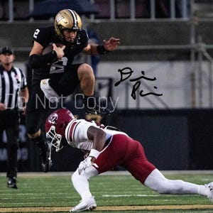 May include: A football player in a black and gold uniform jumps over another player in a maroon and white uniform on a green field. The player in black holds a football. A referee in black and white stripes stands in the background.