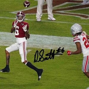 May include: Two football players in action on a green field. One player, wearing a maroon jersey with the number 6, white pants, and a helmet, attempts to catch a football. Another player, in a white and red jersey, runs to the right.