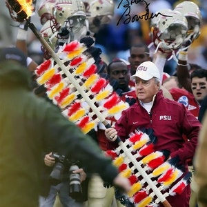 May include: A football coach wearing a maroon jacket with the letters "FSU" on the front, holds a ceremonial spear with red, yellow, and white feathers. The spear is lit on fire at the top. The coach is surrounded by football players wearing helmets.