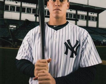 May include: A baseball player wearing a white and black striped jersey with the New York Yankees logo, a black baseball cap with the New York Yankees logo, and a black and white baseball bat. The player is holding the bat in a batting stance. The player's signature is in the bottom right corner of the image.