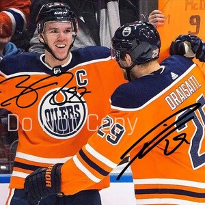 May include: Two hockey players wearing orange and blue jerseys with the team logo and player numbers. The players are signing their names on their jerseys. The jersey on the left has the name "McDavid" and the number "97". The jersey on the right has the name "Draisaitl" and the number "29".