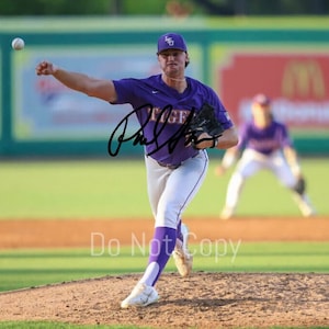 May include: A baseball pitcher in a purple jersey with the word "Tigers" in gold lettering throws a baseball. The pitcher is wearing a purple baseball cap and white pants. The image is taken on a baseball field.