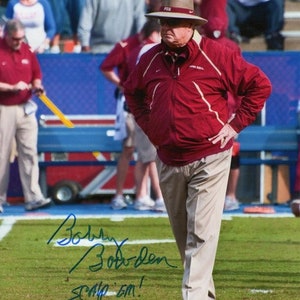 May include: A man wearing a maroon jacket and a brown hat with the letters "FSU" on the front. He is standing on a green field with a blue and white fence in the background. The man is holding his hands on his hips and has a serious expression on his face. The man's signature is written in blue ink below him, which reads "Bobby Bowden,  SCAMP 'EM!"