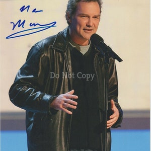 May include: A man wearing a black leather jacket and a black shirt is speaking into a microphone. He is standing on a stage with a blue background. The man's signature is in blue ink in the top left corner of the image. The signature reads "Mark Maron".