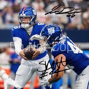 May include: Two football players in blue and white uniforms, one holding a brown football. Both players wear helmets with the New York Giants logo. The image includes signatures. The background is blurred, suggesting a game setting.