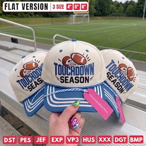 Op de afbeelding: Drie gebroken witte baseballpetten met blauwe kleppen en een blauw en bruin geborduurd ontwerp met twee footballs en de tekst TOUCHDOWN SEASON. De petten bevatten een blauw-wit gedraaid koord en een roze label met DesiraesKitchen.