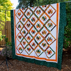 Peut inclure: Une courtepointe faite à la main avec un fond blanc, présentant un motif répété de formes de feuilles colorées en orange, vert et rouge. La courtepointe a une bordure orange et un bord vert à motifs, exposée à l'extérieur.