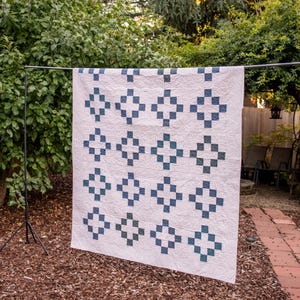 May include: A rectangular quilt featuring a white background with a repeating pattern of dark blue and teal square blocks arranged in a grid. The white fabric displays an intricate, swirling quilted texture across the entire surface area.