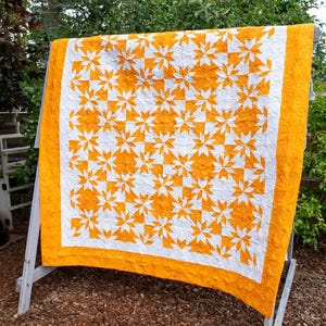 May include: A handmade quilt with a white background and a repeating pattern of orange starbursts. The quilt has an orange border and is displayed outdoors on a wooden drying rack. The quilt is a cozy home decor item.