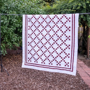 Pode incluir: Uma colcha retangular com fundo branco e um padrão de grade repetitivo de pequenos quadrados florais em vermelho escuro. A colcha tem uma borda larga do mesmo tecido floral vermelho escuro e um design de costura à máquina em espiral.