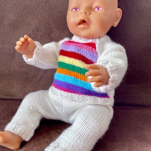 May include: A white knitted baby doll outfit with a rainbow striped pattern on the chest. The doll is sitting on a brown surface.