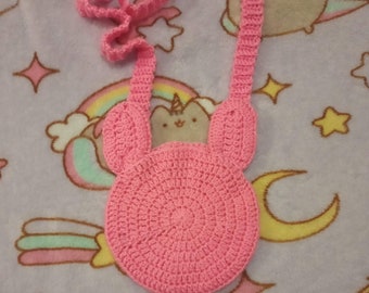 May include: A pink crocheted shoulder bag with a round body and two ear-shaped flaps. The bag has a long strap.