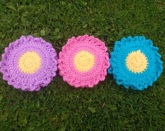 Könnte beinhalten: Drei gehäkelte Untersetzer in Form von Blumen in Lila, Rosa und Blau mit gelben Zentren.