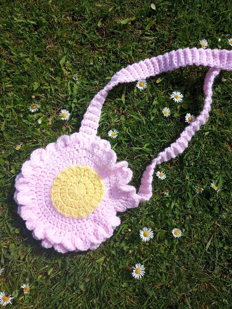 May include: A pink crocheted flower-shaped bag with a yellow center. The bag has a long pink crocheted strap. The bag is lying on a bed of green grass with white daisies.