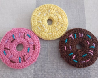 May include: Three crocheted donuts, one pink with brown and blue sprinkles, one yellow with white sprinkles, and one brown with pink and blue sprinkles.