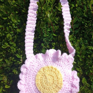 May include: A pink crocheted flower-shaped bag with a yellow center. The bag has a long pink crocheted strap.