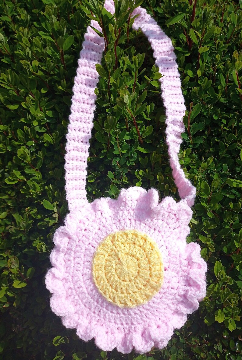 May include: A pink crocheted flower-shaped bag with a yellow center. The bag has a long pink crocheted strap.