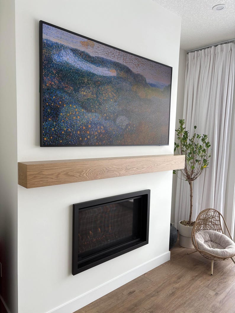 May include: A modern living room featuring a large, framed artwork with a colorful landscape scene above a wooden mantel. Below, a black-framed fireplace is set into a white wall. A wicker chair and a potted plant add to the room's decor.