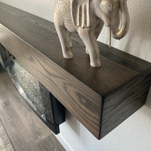 May include: A decorative gray elephant figurine with gold accents stands on a dark brown wooden mantel. The mantel is mounted on a white wall. The elephant has detailed carvings.