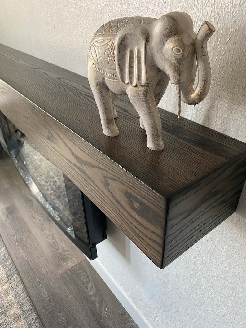 May include: A decorative gray elephant figurine with gold accents stands on a dark brown wooden mantel. The mantel is mounted on a white wall. The elephant has detailed carvings.