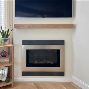 May include: A modern fireplace with a stainless steel frame and a black border. Above it is a wooden shelf, and a black television is mounted on the wall. The room has a minimalist design.