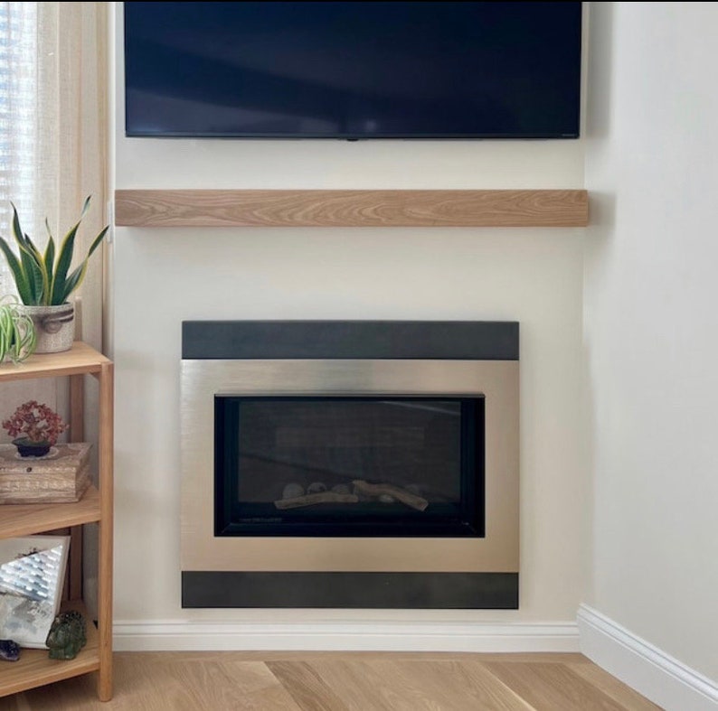 May include: A modern fireplace with a stainless steel frame and a black border. Above it is a wooden shelf, and a black television is mounted on the wall. The room has a minimalist design.