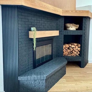 May include: A modern black brick fireplace with a wooden mantel and built-in storage. The fireplace features a glass door and a bench. The storage area holds firewood and a decorative basket. The design incorporates wood and black brick.