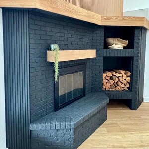 May include: A modern black brick fireplace with a wooden mantel and built-in storage. The fireplace features a glass door and a bench. The storage area holds firewood and a decorative basket. The design incorporates wood and black brick.