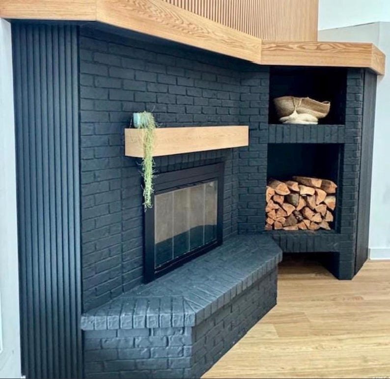 May include: A modern black brick fireplace with a wooden mantel and built-in storage. The fireplace features a glass door and a bench. The storage area holds firewood and a decorative basket. The design incorporates wood and black brick.