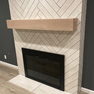 May include: A fireplace with a light-colored herringbone tile surround. It features a light wood mantel and a black framed firebox with glass doors. The hearth is made of matching tile, and the wall is painted a dark gray.