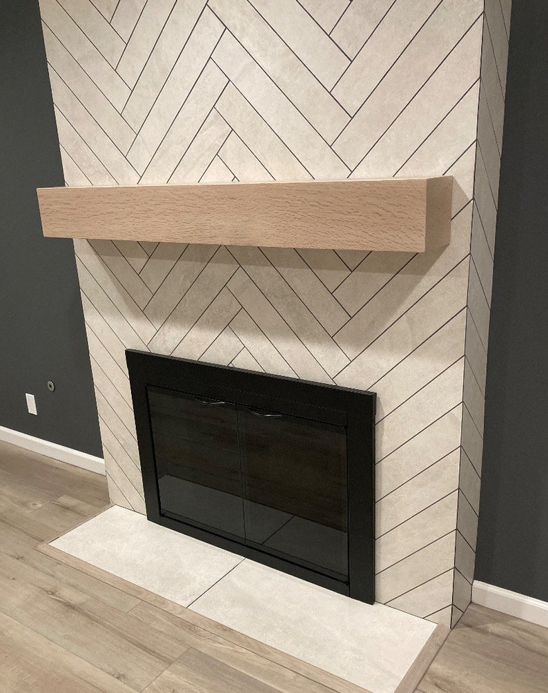 May include: A fireplace with a light-colored herringbone tile surround. It features a light wood mantel and a black framed firebox with glass doors. The hearth is made of matching tile, and the wall is painted a dark gray.