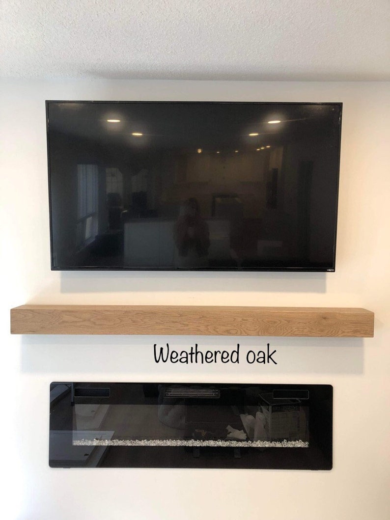 May include: A large black television mounted on a white wall, above a weathered oak floating shelf. Below, a black electric fireplace with a glass front and decorative crystals. The text "Weathered oak" is below the shelf.