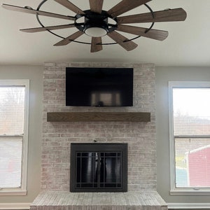 May include: A modern living room featuring a brick fireplace with a mounted television and a wooden mantel. Above, a ceiling fan with wooden blades and a black frame adds a stylish touch. The room is illuminated by natural light from the windows.
