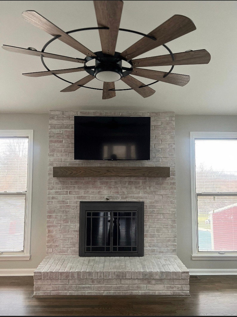 May include: A modern living room featuring a brick fireplace with a mounted television and a wooden mantel. Above, a ceiling fan with wooden blades and a black frame adds a stylish touch. The room is illuminated by natural light from the windows.