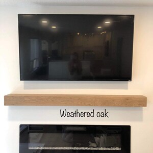 May include: A large black flat-screen television mounted on a white wall, above a weathered oak floating shelf. Below the shelf is a black electric fireplace with a glass front and decorative stones. The text "Weathered oak" is below the shelf.