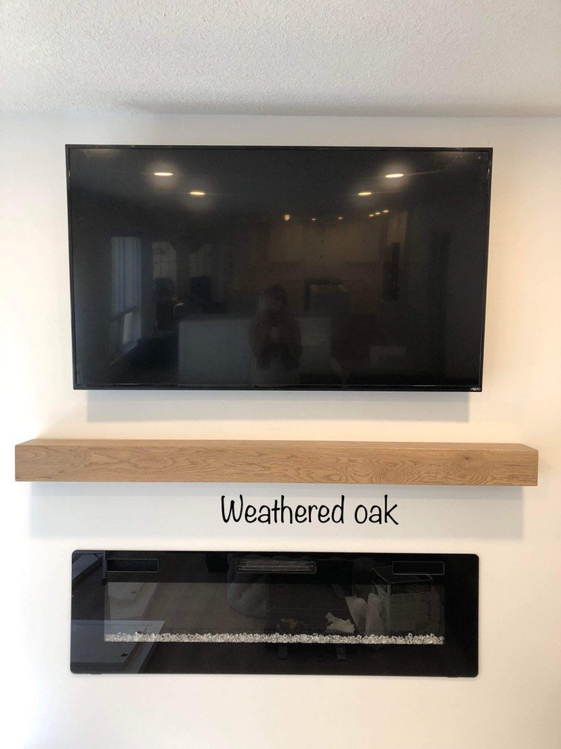 May include: A large black flat-screen television mounted on a white wall, above a weathered oak floating shelf. Below the shelf is a black electric fireplace with a glass front and decorative stones. The text "Weathered oak" is below the shelf.
