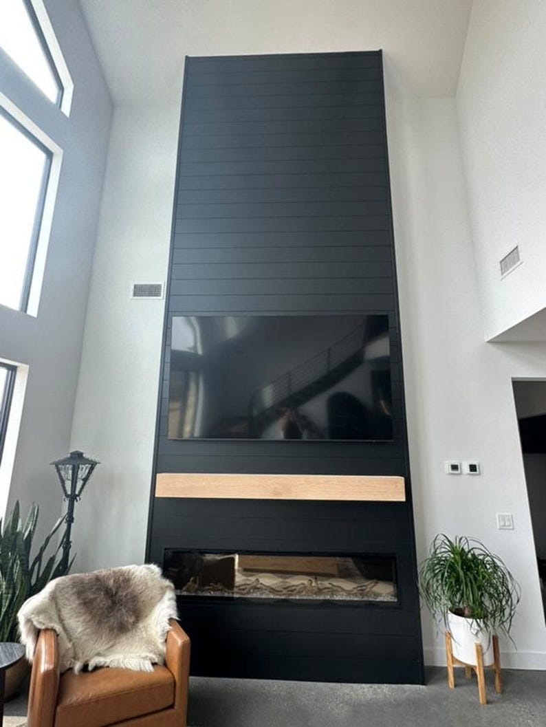 May include: A modern black fireplace with a built-in television and a light wood mantel. The fireplace has a linear design and is flanked by a leather armchair with a fur throw and a potted plant.