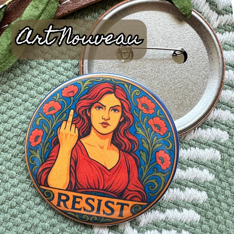 Op de afbeelding: Een ronde button met een Art Nouveau-ontwerp. De button toont een vrouw met rood haar en een rode jurk, die de middelvinger opsteekt. Het woord "RESIST" staat onderaan. De achtergrond is blauw met rode bloemen.