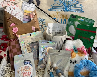 May include: A gift box containing a canvas tote bag with a blue floral book graphic, a mug labeled Instant Hot Cocoa Mix, a brown envelope with bookmarks, a checkered notebook, a sticker reading Be Proud Of Who You Are, a beaded keychain, fuzzy socks, and face masks.