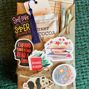 May include: A brown paper envelope filled with reading-themed items. Includes stickers with phrases like "Self Love is Your Super Power" and "Expand Your Mind, Read a Book." Also features a reading log, cocoa packet, and a blue tassel.