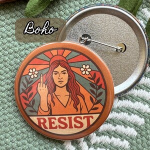 Op de afbeelding: Een ronde button met een retro design. Het toont een vrouw die de middelvinger opsteekt, met het woord "RESIST" onderaan. De button heeft een speld aan de achterkant en het woord "Boho" staat in de linkerbovenhoek.