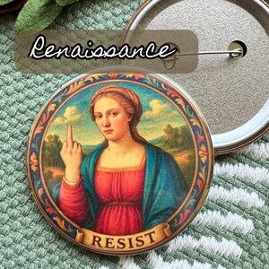 Op de afbeelding: Ronde knoopspeld met een afbeelding in renaissancestijl van een vrouw die de middelvinger opsteekt. De afbeelding is omlijst met een decoratieve rand en het woord "RESIST" staat op een banner. Het woord "Renaissance" staat boven de knop.