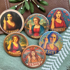 May include: A collection of five round buttons featuring Renaissance-style portraits of women giving the middle finger. Each button has the word "RESIST" printed on it. The buttons are in various colors, including red, blue, and brown.