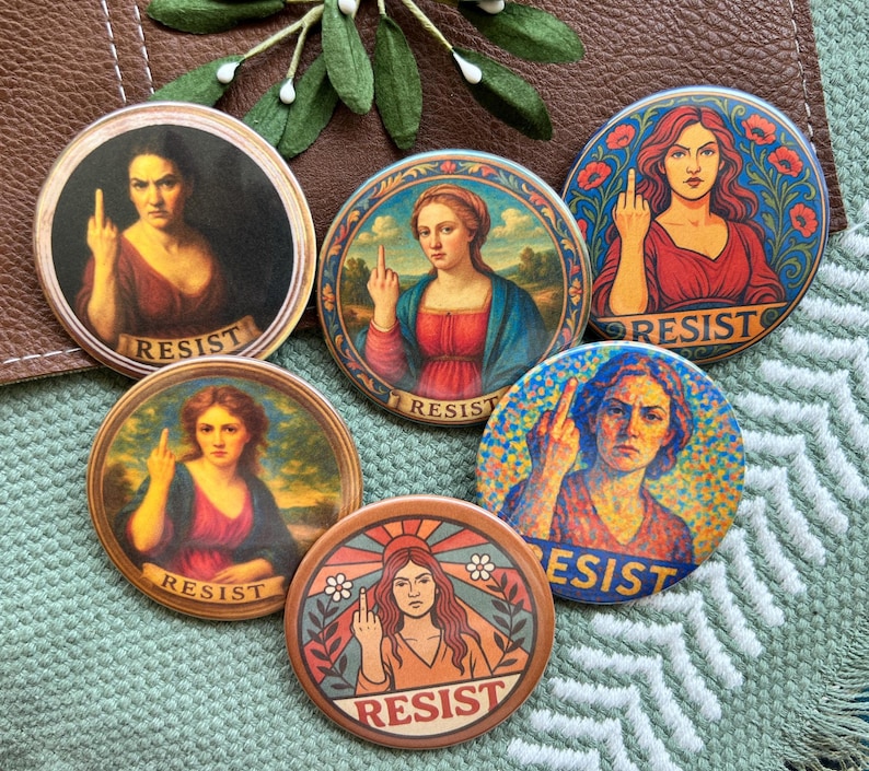 Op de afbeelding: Een verzameling van vijf ronde buttons met portretten in renaissancestijl van vrouwen die de middelvinger opsteken. Op elke button staat het woord "RESIST" gedrukt. De buttons zijn in verschillende kleuren, waaronder rood, blauw en bruin.