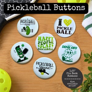 May include: Five round pin-back buttons featuring pickleball-themed graphics and text. Designs include a pickle character, a heart, and phrases like Pickleball Crew, I Love Pickleball, Happy People Play Pickleball, Here to Dink Off, and Pickleball Squad.