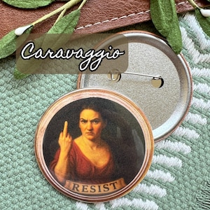 Op de afbeelding: Een ronde button met een schilderij van een vrouw die de middelvinger opsteekt, met het woord "RESIST" op een spandoek. De button heeft een metalen pin en het woord "Caravaggio" staat erboven.