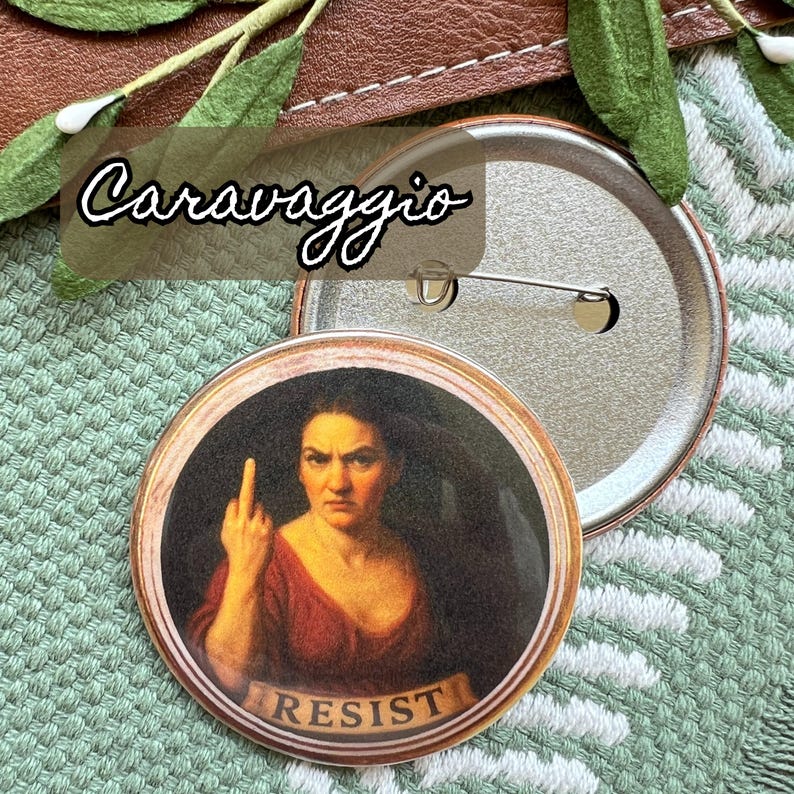 Op de afbeelding: Een ronde button met een schilderij van een vrouw die de middelvinger opsteekt, met het woord "RESIST" op een spandoek. De button heeft een metalen pin en het woord "Caravaggio" staat erboven.