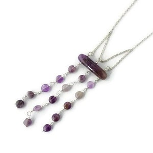 Op de afbeelding: Een zilveren ketting met een lange, rechthoekige amethist hanger en meerdere strengen kleinere amethist kralen die aan de hanger hangen.