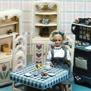 May include: A miniature dollhouse kitchen set crafted from plastic canvas. The set features a cabinet, corner cabinet, stove, table, chairs, and rug. The furniture is white, blue, and brown with floral and heart patterns. A doll is seated at the table.