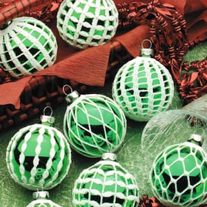 May include: A collection of seven green glass Christmas baubles with white crocheted covers. The baubles are arranged in a basket and on a green surface.